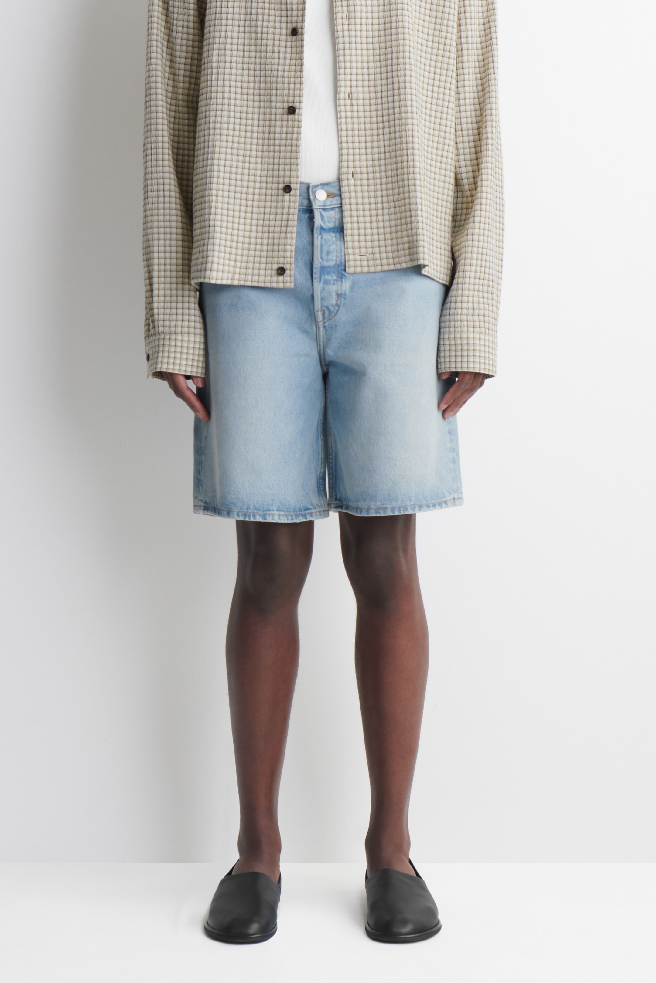 Person wearing a checkered beige shirt, light blue denim shorts, and black slip-on shoes, standing against a white background.
