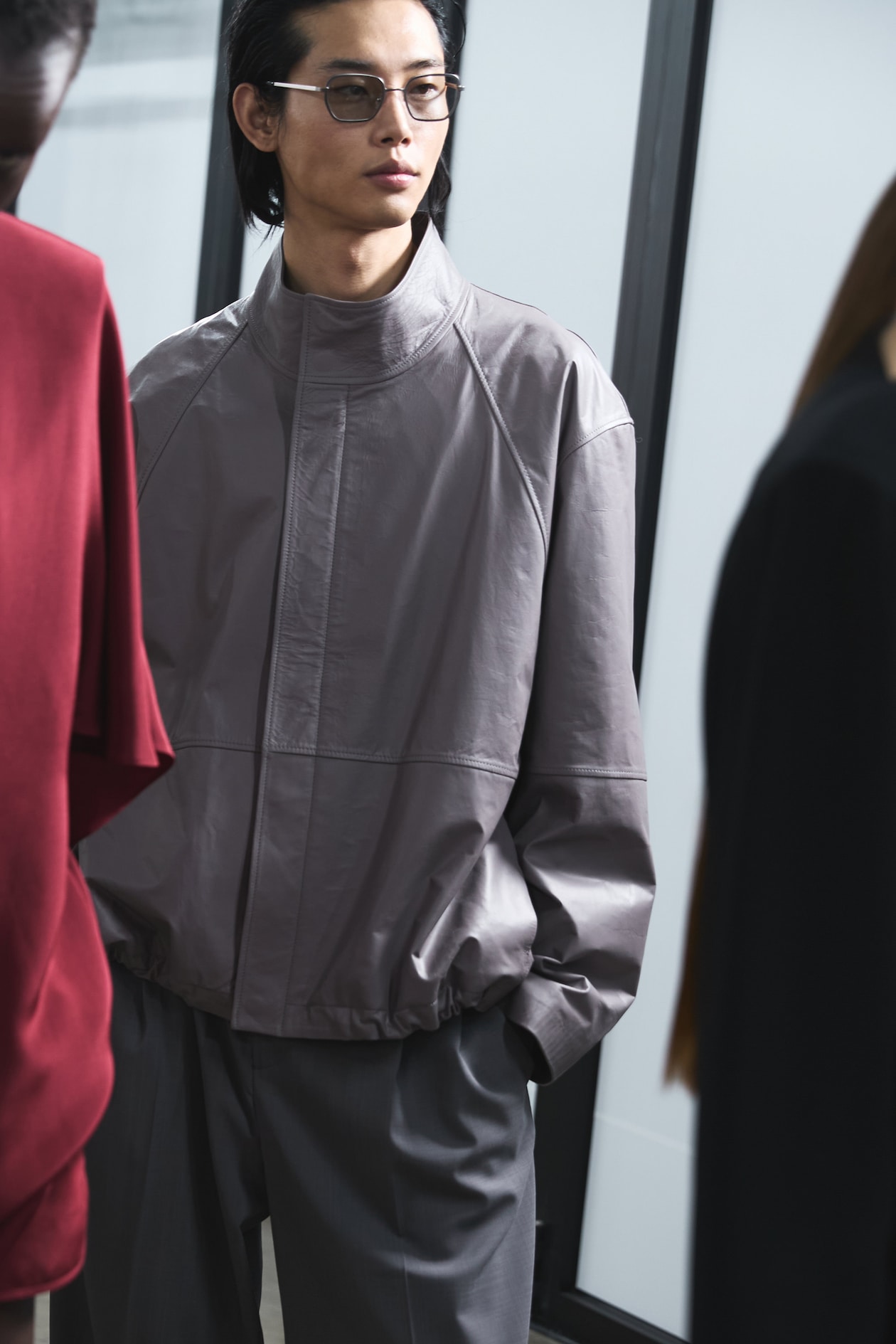 Person wearing glasses and a light gray jacket, standing indoors with others nearby, looking to the side.