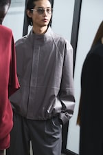 Placeholder for: Person wearing glasses and a light gray jacket, standing indoors with others nearby, looking to the side.