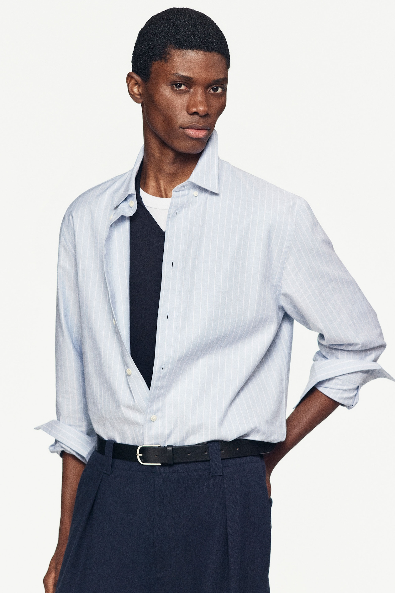 Person wearing a light blue striped shirt over a black shirt, with dark pants and a belt, posing against a plain white background.