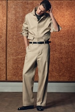 Placeholder for: Person in a beige jacket and pants, with a black belt and shoes, stands against a textured brown wall, looking down with one hand on their head.