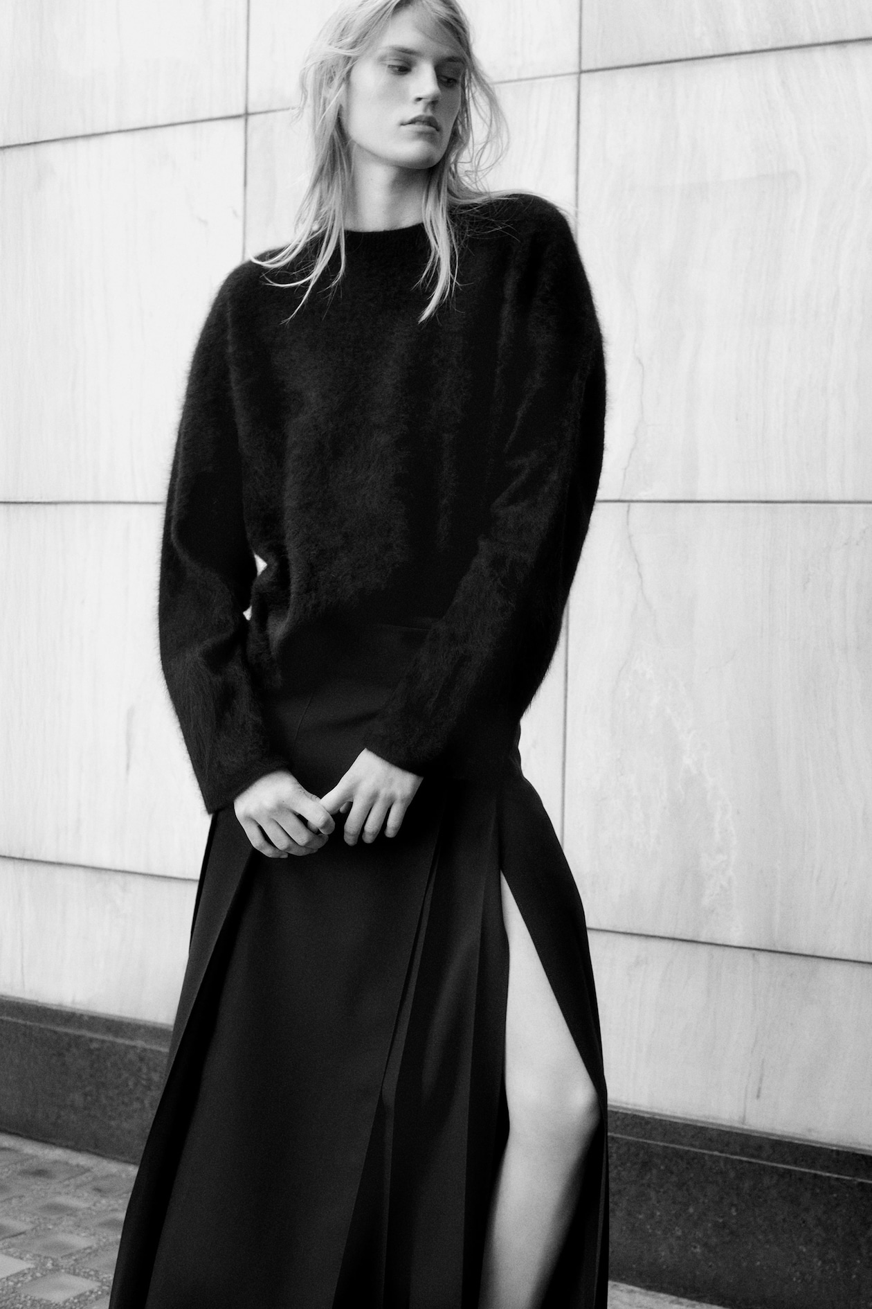 Person in a black fuzzy sweater and long skirt with a slit, standing against a marble wall, gazing forward. Black and white image.