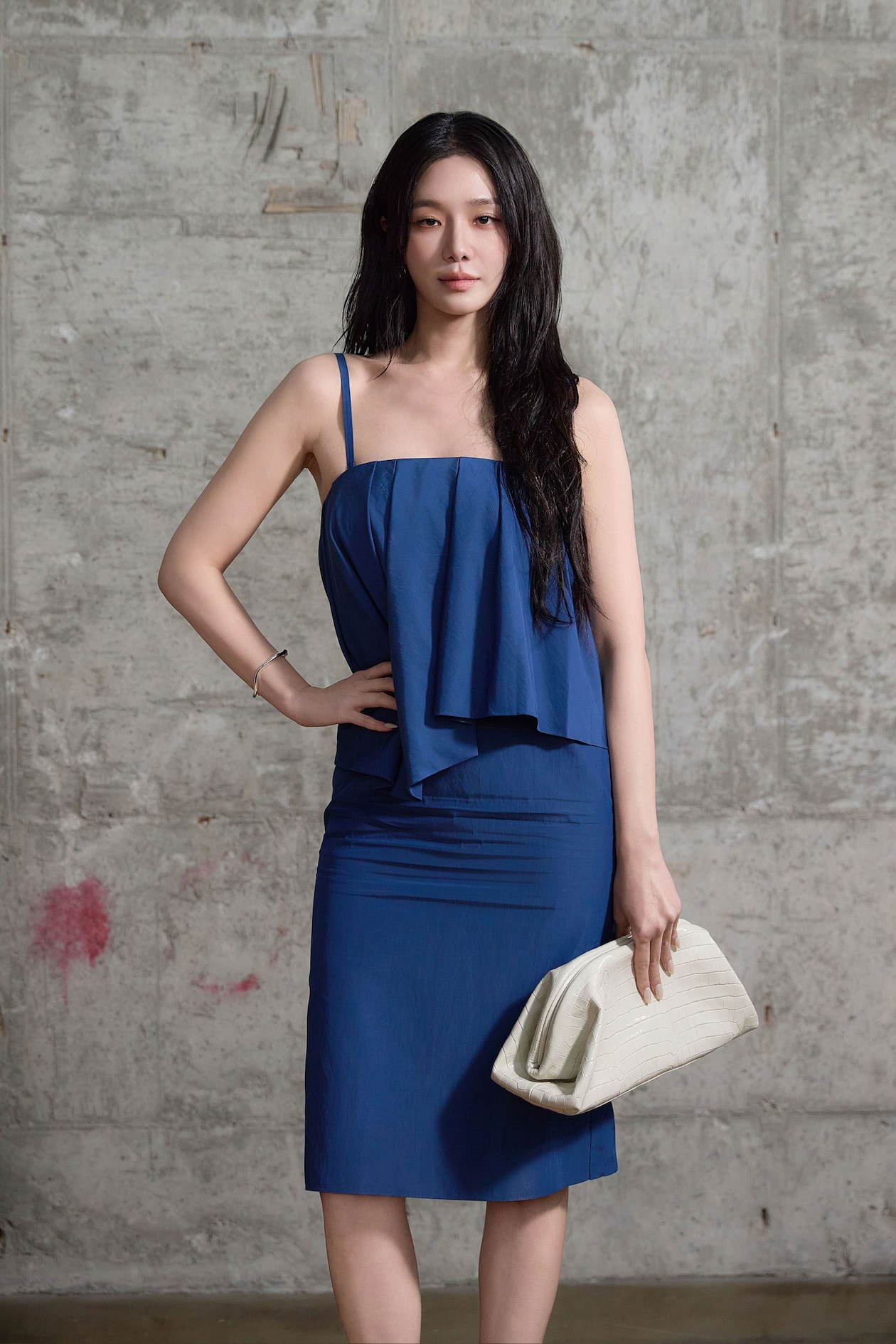 Woman in a blue dress stands confidently against a textured gray wall, holding a white clutch.