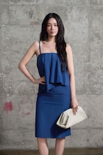 Placeholder for: Woman in a blue dress stands confidently against a textured gray wall, holding a white clutch.