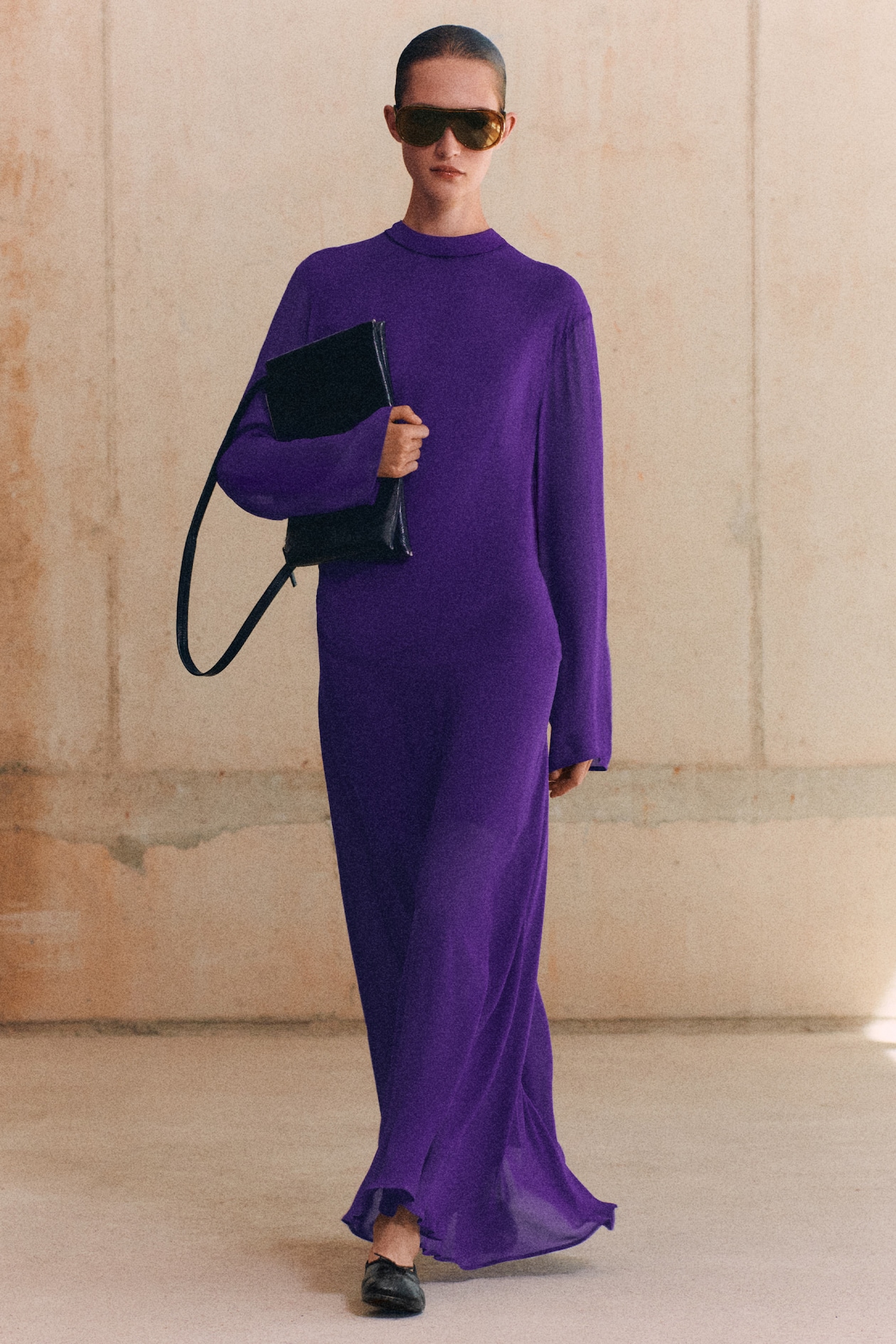 Person wearing a long purple dress, sunglasses, and black shoes, holding a black purse, standing against a beige wall.