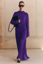 Placeholder for: Person wearing a long purple dress, sunglasses, and black shoes, holding a black purse, standing against a beige wall.