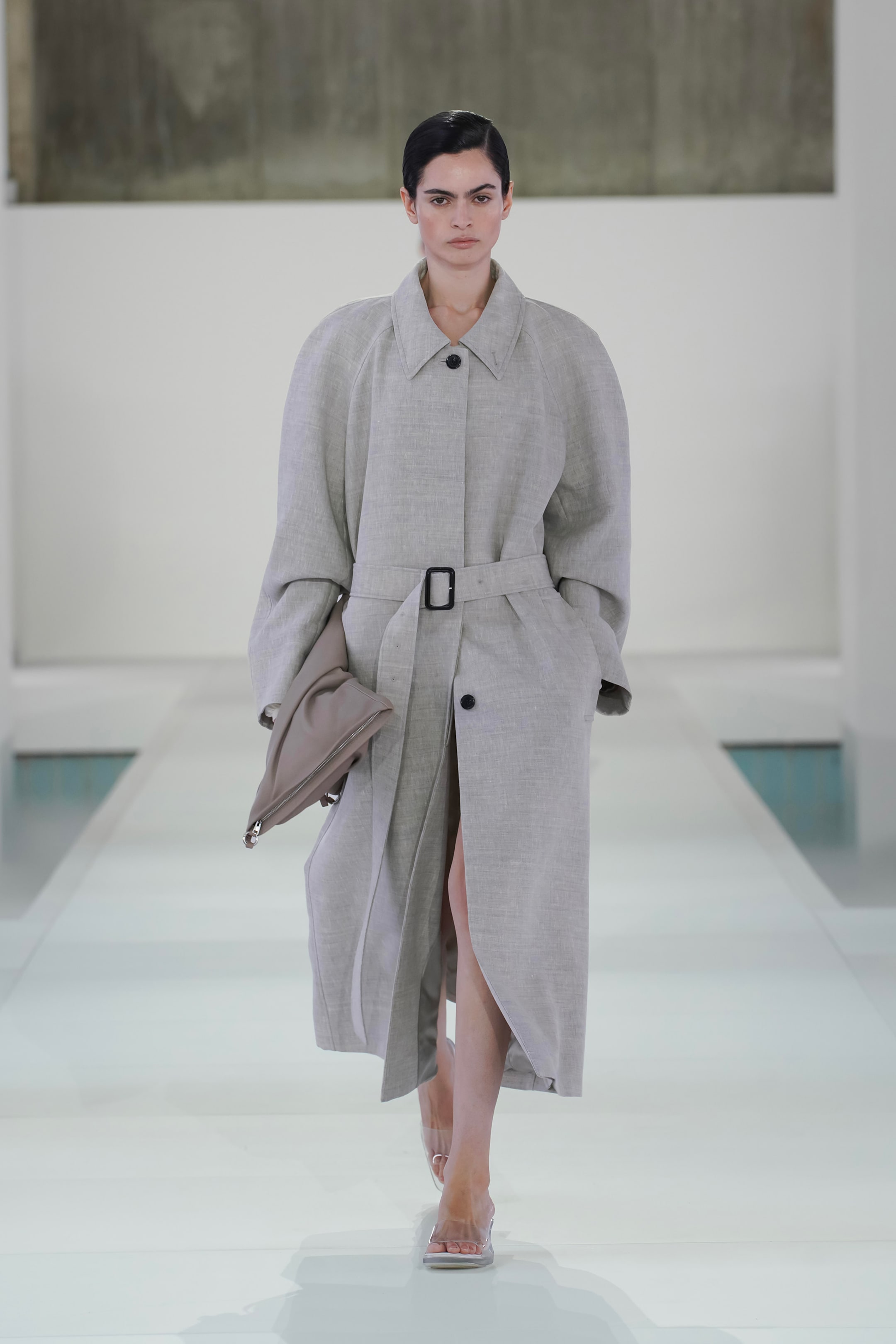 A model in a long, belted grey coat and clear heels walks down a white runway with a neutral background.