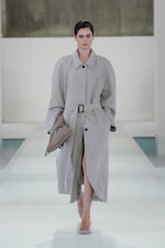 Placeholder for: A model in a long, belted grey coat and clear heels walks down a white runway with a neutral background.