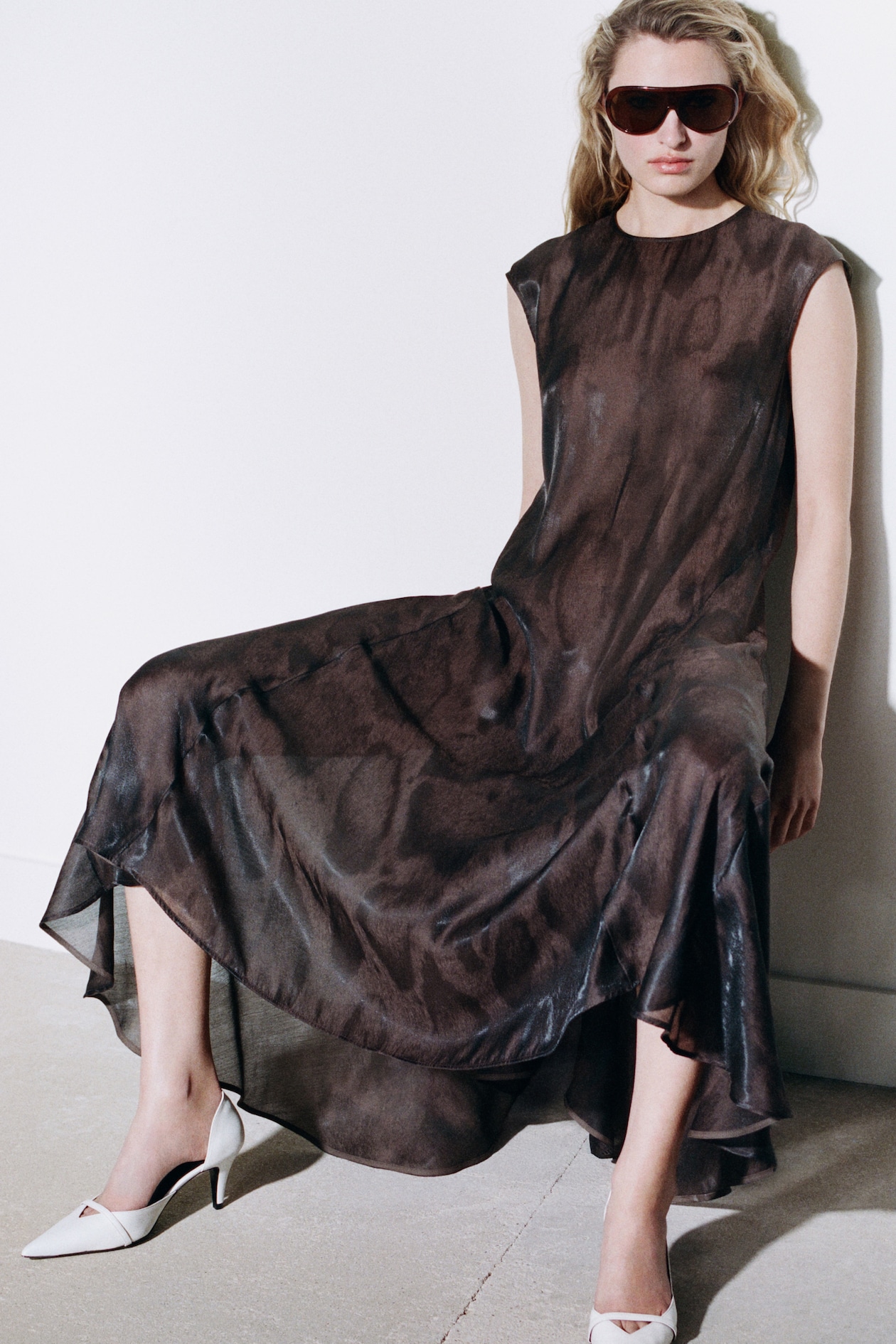 Woman in a flowing, dark sheer dress and white heels, wearing large sunglasses, leans against a white wall on a concrete floor.