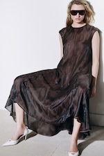 Placeholder for: Woman in a flowing, dark sheer dress and white heels, wearing large sunglasses, leans against a white wall on a concrete floor.