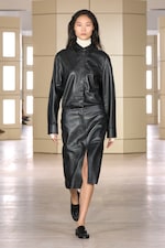 Placeholder for: Model wearing a black leather outfit with a slit skirt and white turtleneck, walking on a minimalist indoor runway.