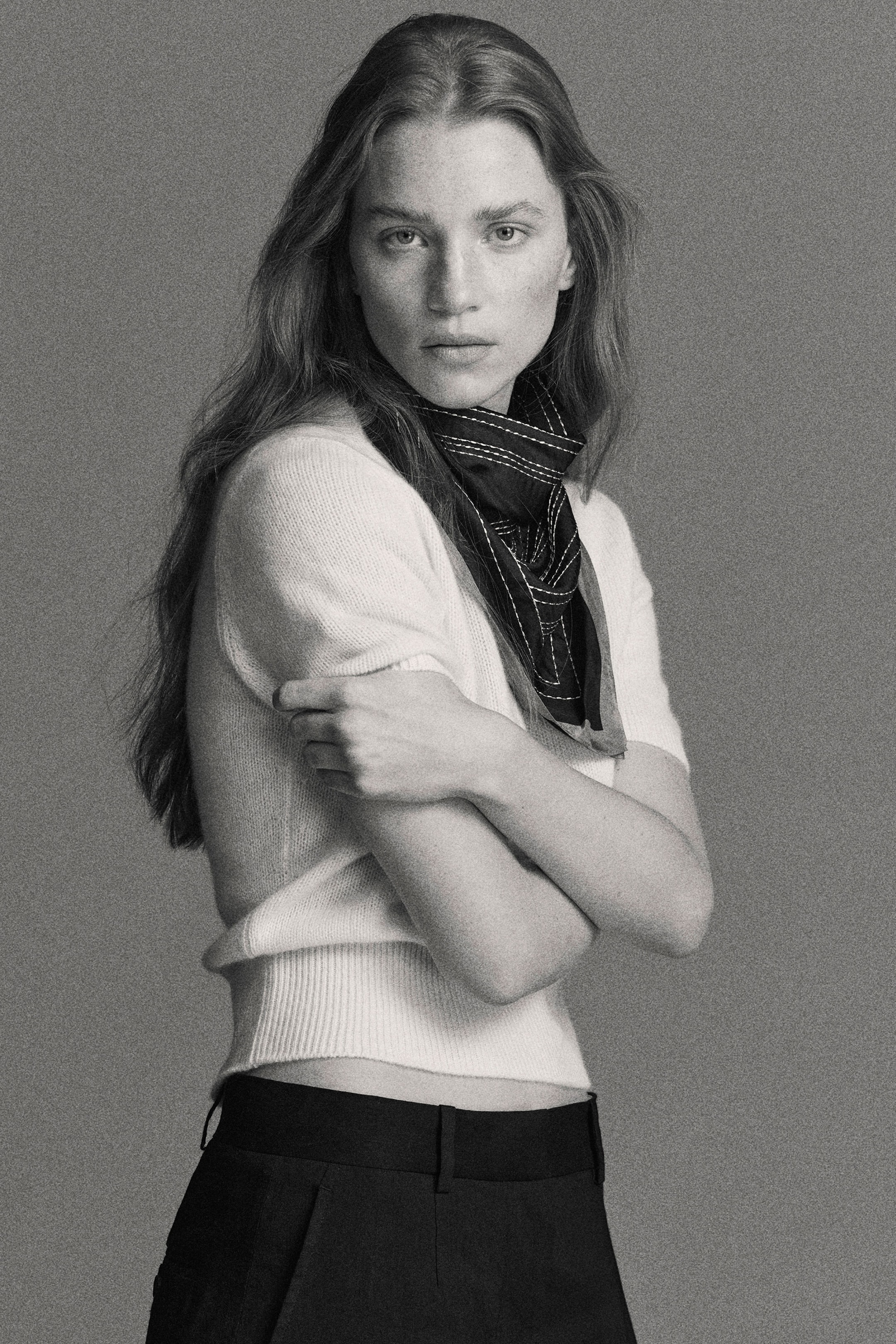 Black and white photo of a person with long hair, wearing a short-sleeve top, scarf, and pants, posing with folded arms against a plain backdrop.