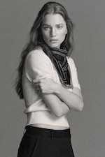 Placeholder for: Black and white photo of a person with long hair, wearing a short-sleeve top, scarf, and pants, posing with folded arms against a plain backdrop.