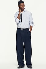 Placeholder for: Person wearing a light blue striped shirt over a black shirt, with dark pants and a belt, posing against a plain white background.