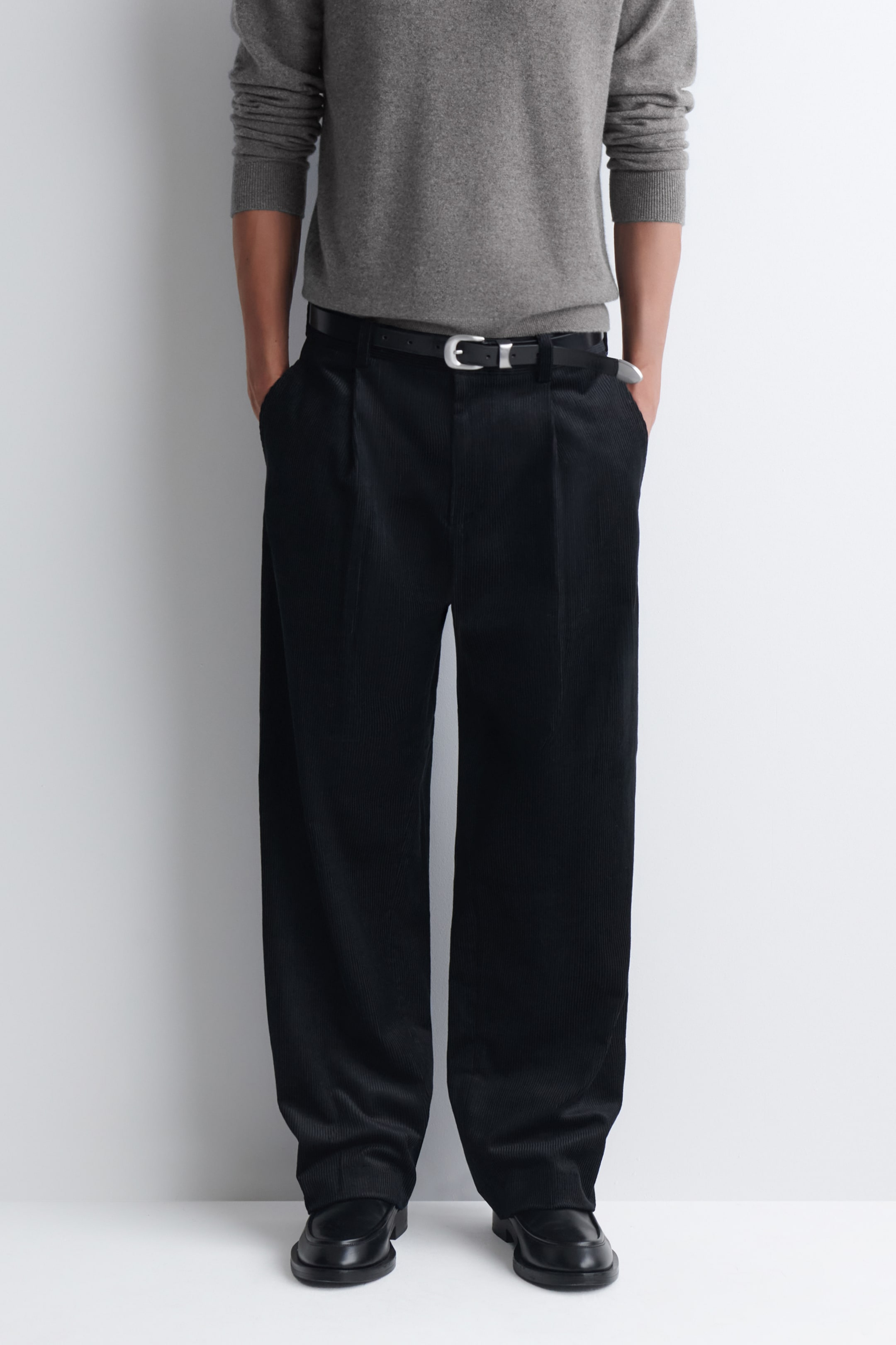 Person wearing a gray sweater, black pleated pants, and black shoes, with hands in pockets against a plain background.