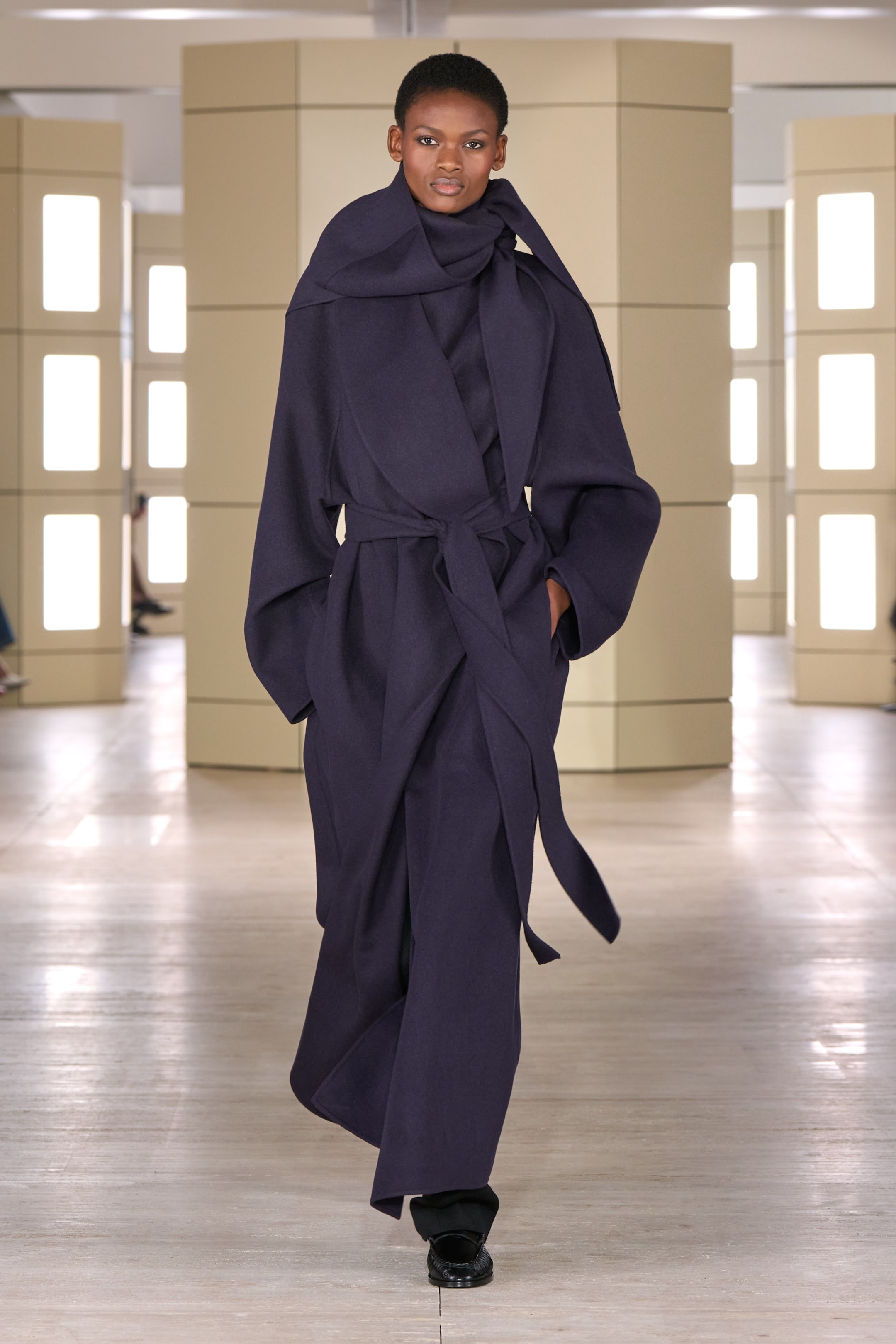 A model walks down a runway wearing a dark purple, long, belted coat with wide lapels, and matching pants, in a bright modern indoor setting.