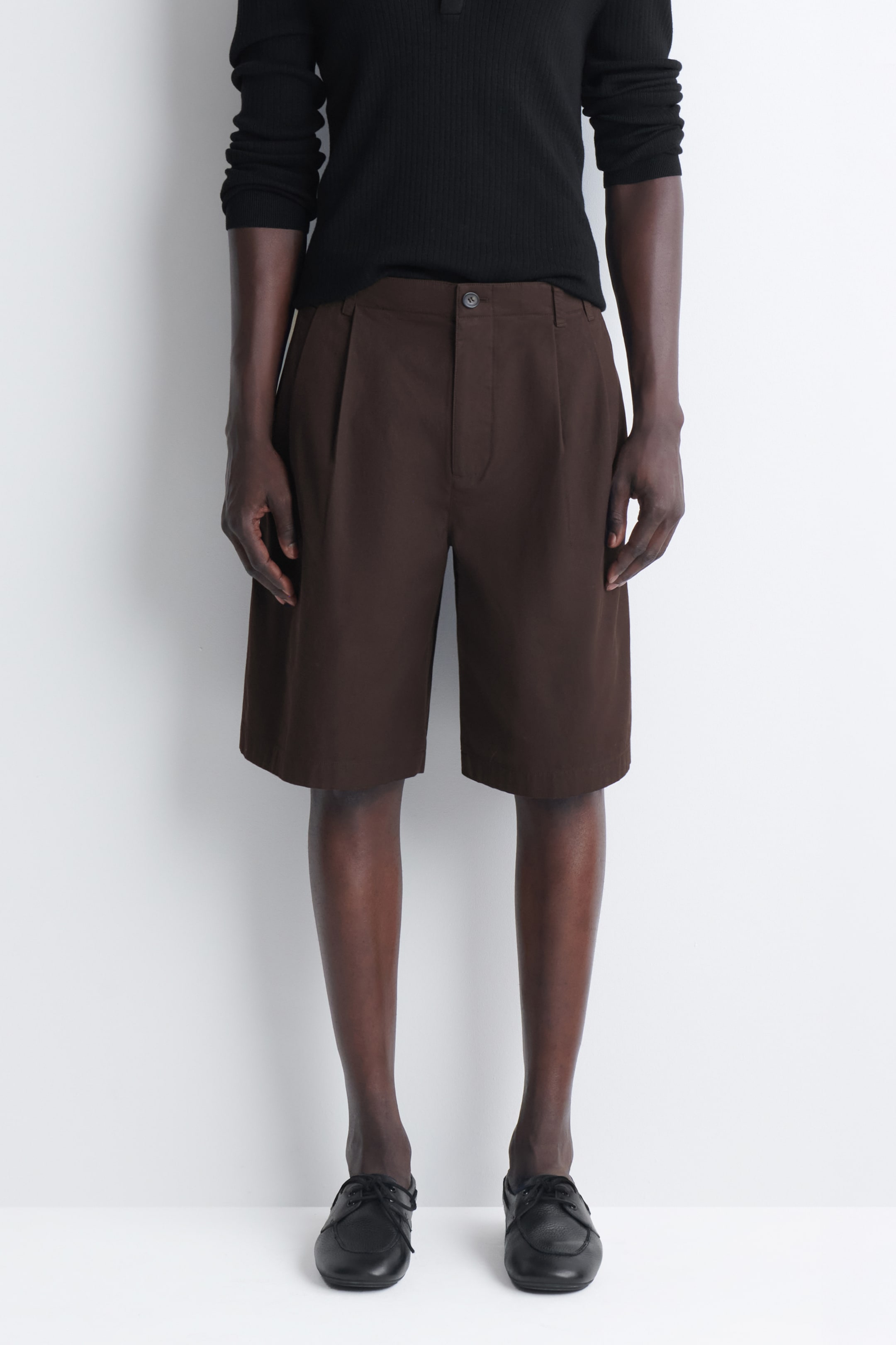 A person stands against a light background wearing a black long-sleeve shirt, brown knee-length shorts, and black shoes.