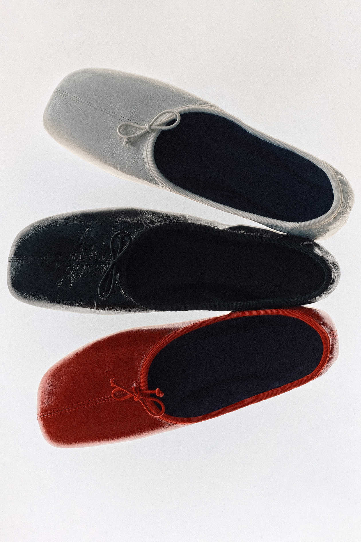 Three pairs of leather slip-on shoes in white, black, and red, each with a small bow, arranged in a vertical stack on a white background.