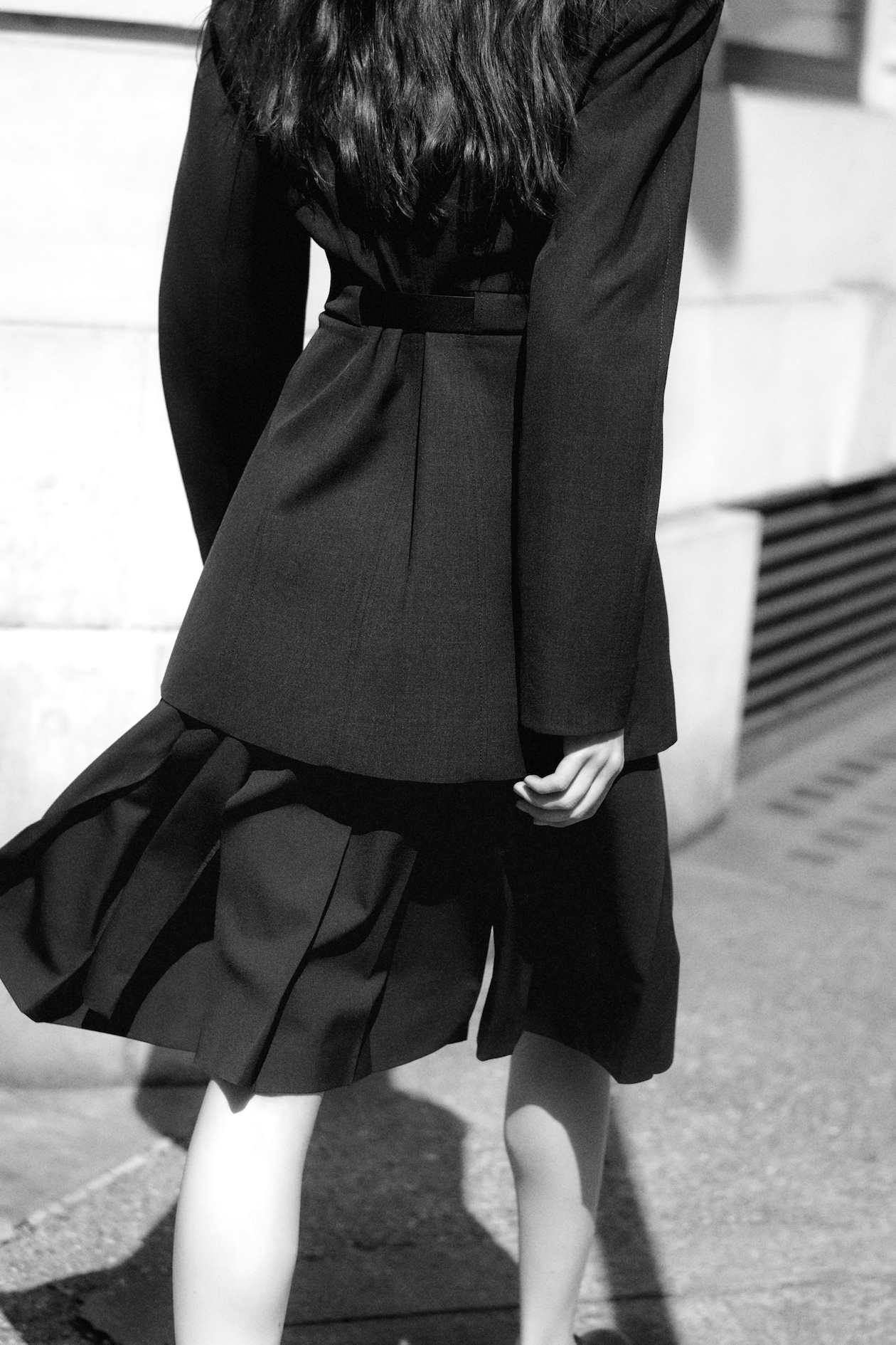 A woman in a dark, tailored outfit walks on a sunlit sidewalk. Her long hair flows down her back, and her skirt catches a slight breeze.