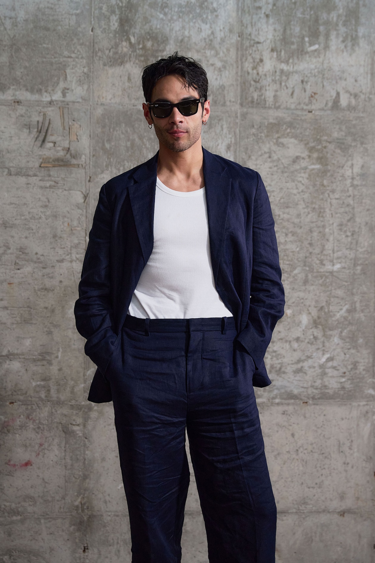 A man in a navy suit and white shirt, wearing sunglasses, stands against a textured concrete wall with hands in his pockets.