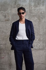 Placeholder for: A man in a navy suit and white shirt, wearing sunglasses, stands against a textured concrete wall with hands in his pockets.