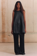 Placeholder for: A woman stands wearing a sleeveless black leather top and black wide-leg pants, with long wavy hair, against a plain background.