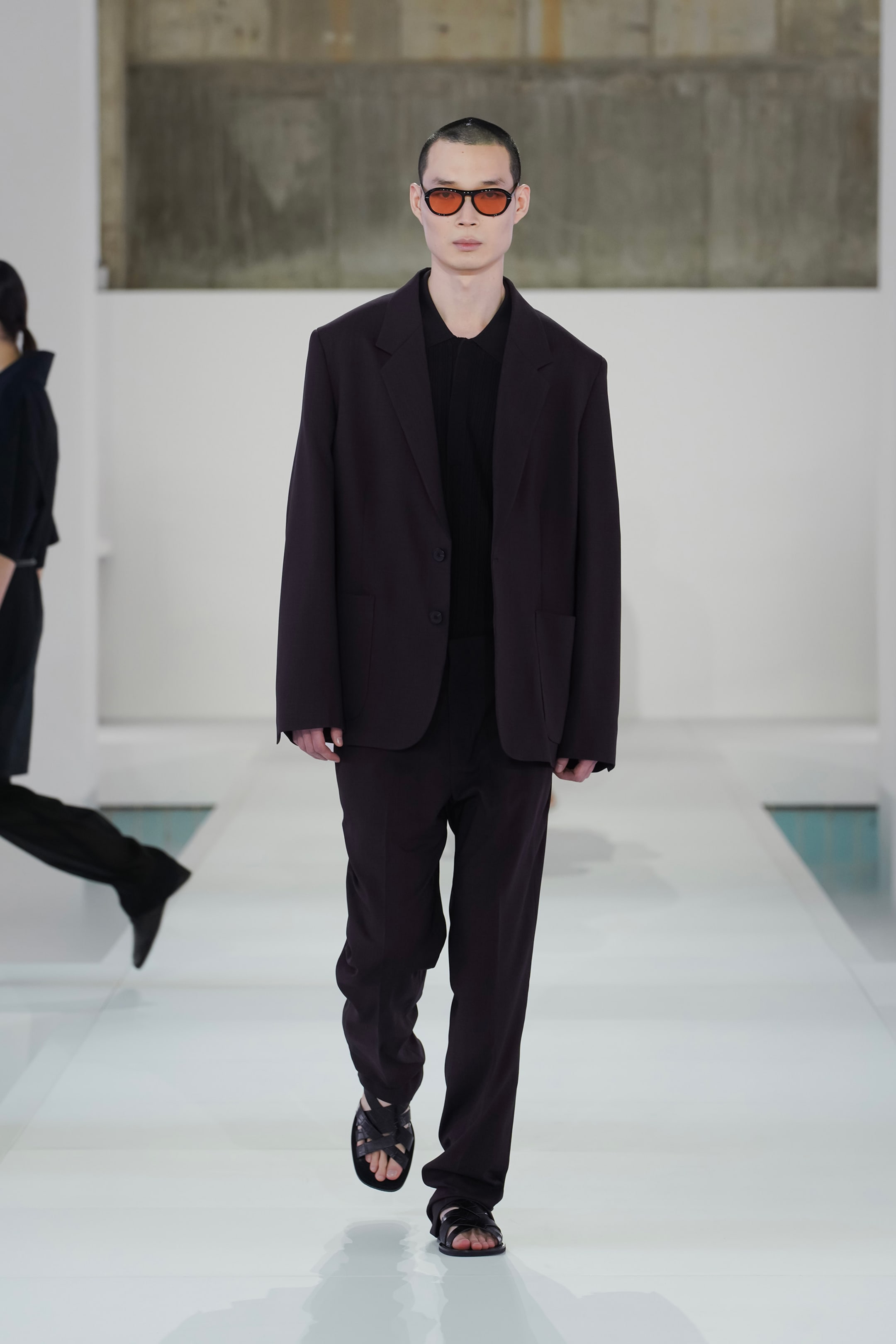 Model in a dark oversized suit and sunglasses walks down a minimalist runway in a fashion show.