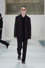 Placeholder for: Model in a dark oversized suit and sunglasses walks down a minimalist runway in a fashion show.