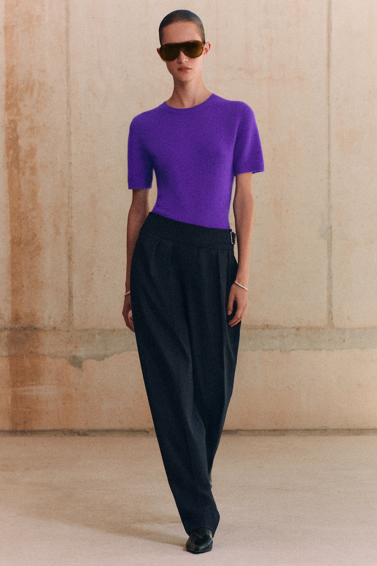 Person wearing a purple top, black wide-leg pants, sunglasses, and bracelets, standing against a beige textured wall.