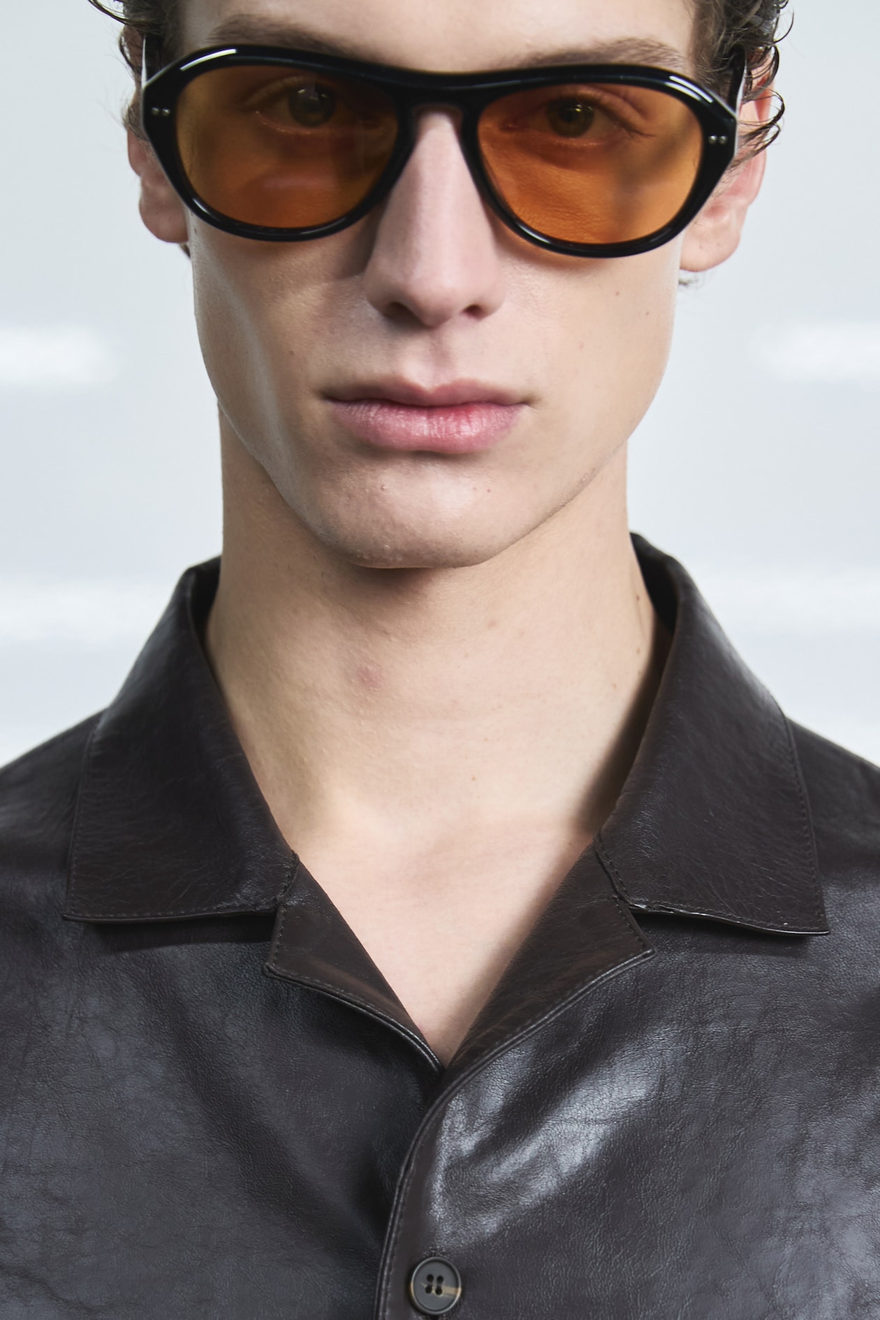 Person wearing oversized brown sunglasses and a dark leather shirt, posing against a light background.