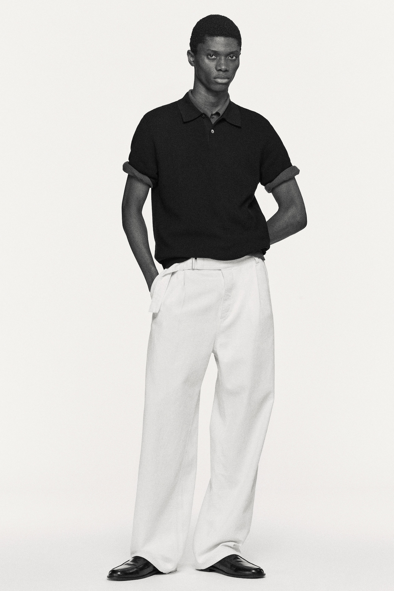 Person in a black short-sleeve shirt and white wide-leg pants, hands in pockets, standing against a plain background.