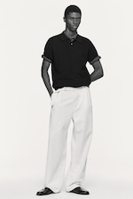 Placeholder for: Person in a black short-sleeve shirt and white wide-leg pants, hands in pockets, standing against a plain background.