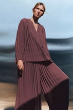 Placeholder for: Person in a pleated, burgundy outfit poses outdoors against a blurred blue and gray background, with sunlight casting shadows.