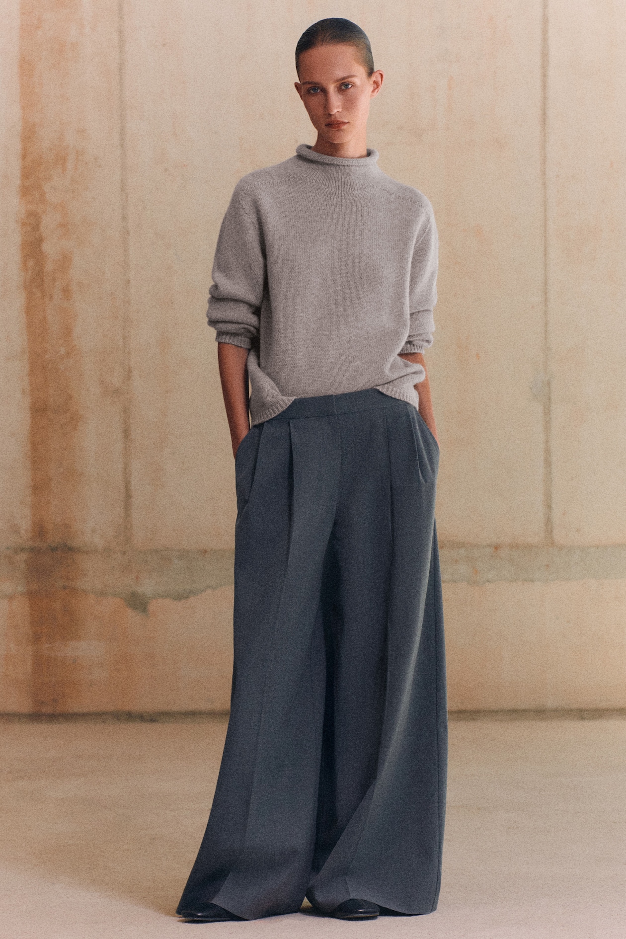 Person wearing a gray sweater and wide-leg gray trousers, standing in a minimalist indoor setting with hands in pockets.