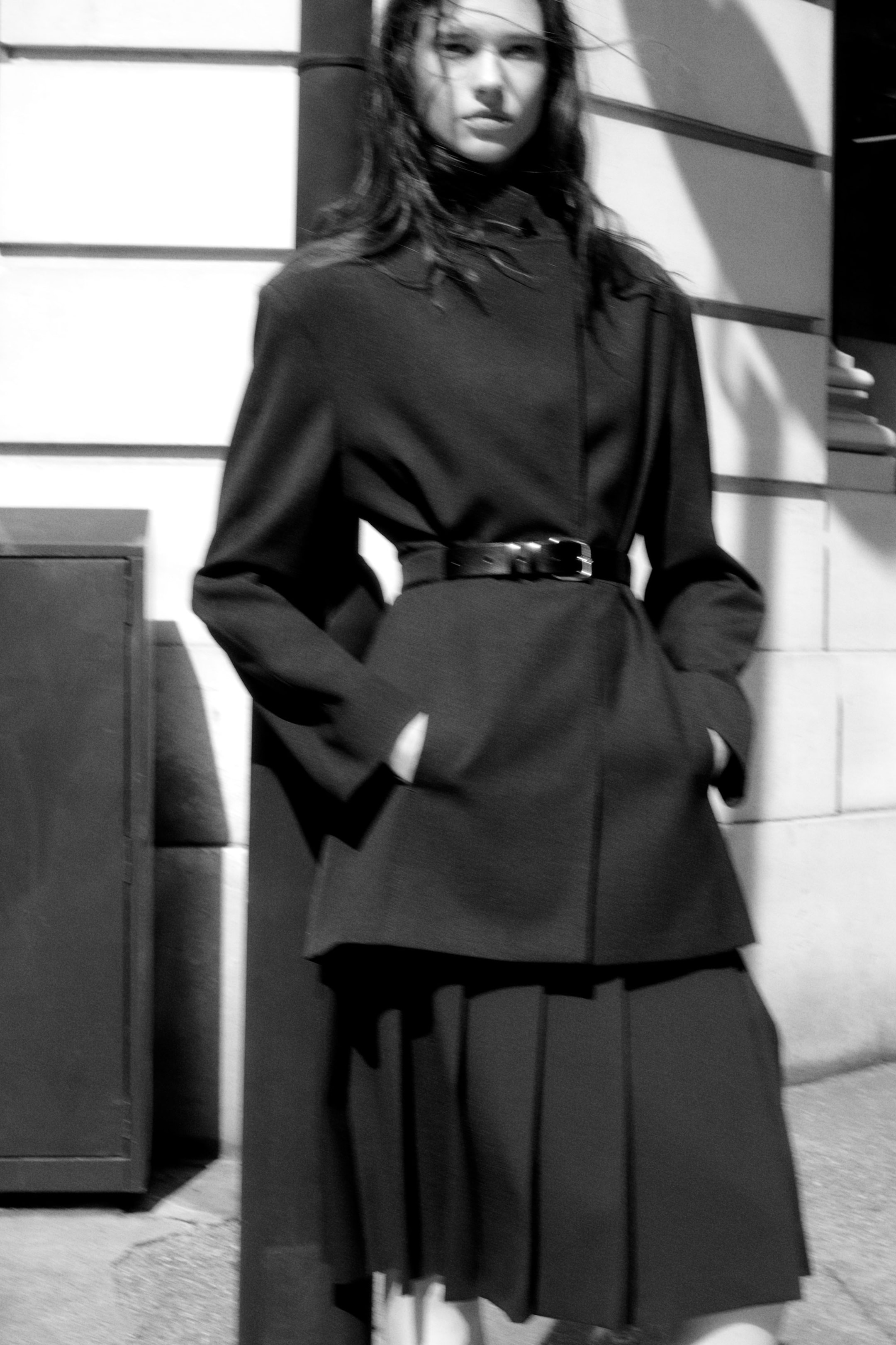 Black and white image of a person in a stylish, layered outfit with a belt, standing against a building at night.