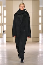 Placeholder for: Person walking on a runway wearing a black double-breasted coat, black pants, and scarf in a brightly lit room with beige panels.