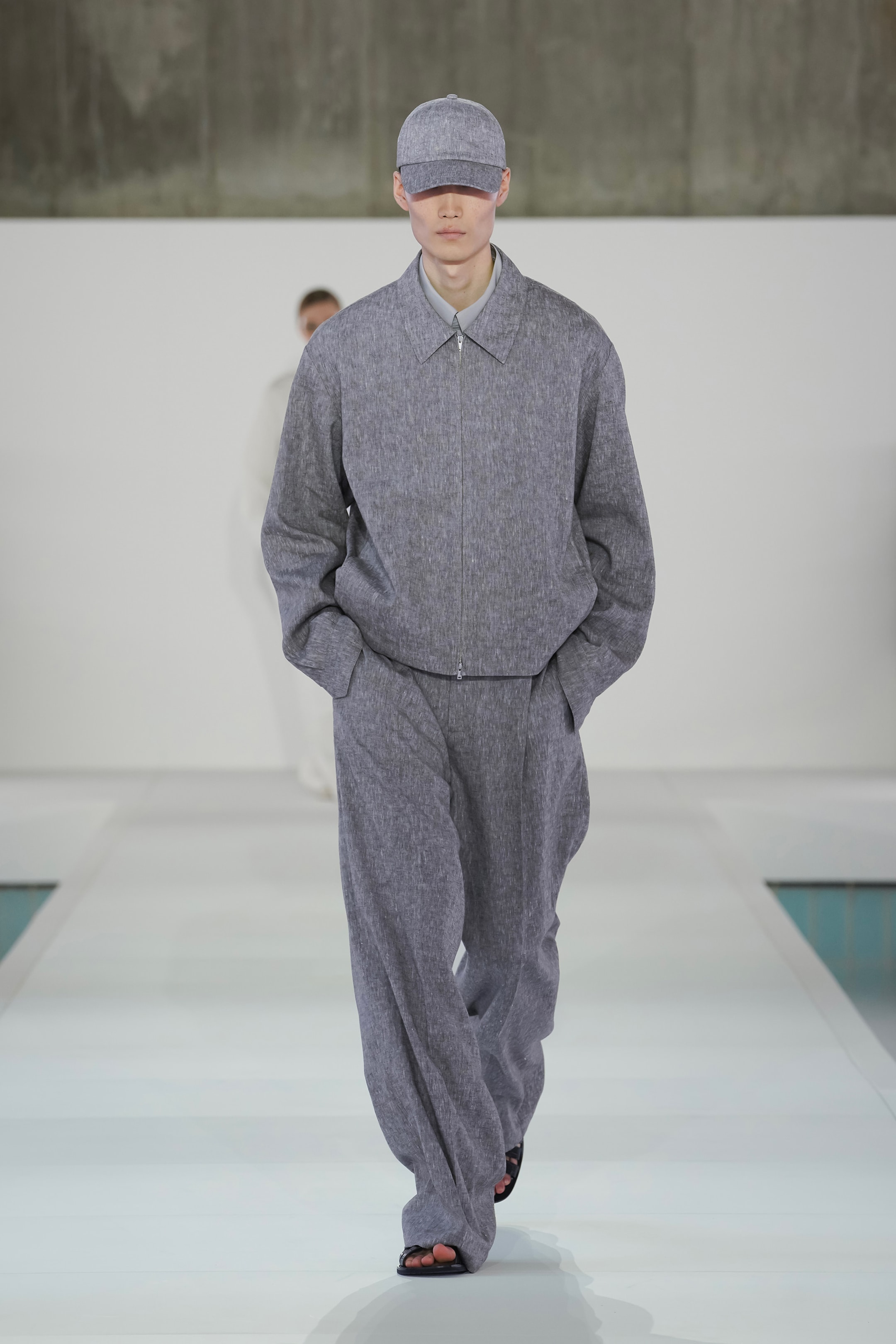 Model in a gray oversized suit and cap walks down a bright runway with a minimalist background.