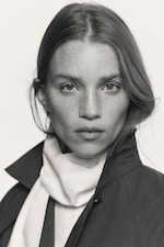 Placeholder for: Black and white portrait of a person wearing a coat and scarf, with long hair and a serious expression.