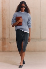 Placeholder for: A woman in sunglasses and a blue sweater holds a red clutch, wearing a black slit skirt and flats, walking indoors against a neutral backdrop.