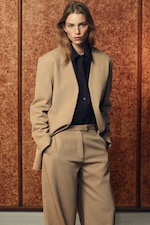 Placeholder for: Person wearing a beige suit and dark shirt stands confidently against a textured brown background.