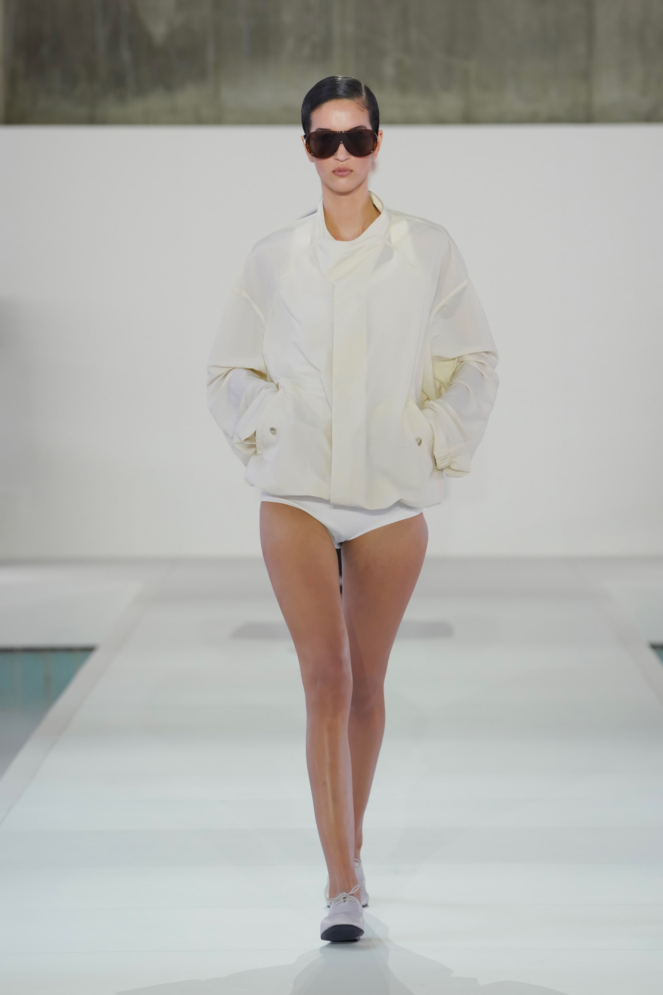 Model walking on a runway wearing a white jacket, white briefs, sunglasses, and heels against a minimalist backdrop.
