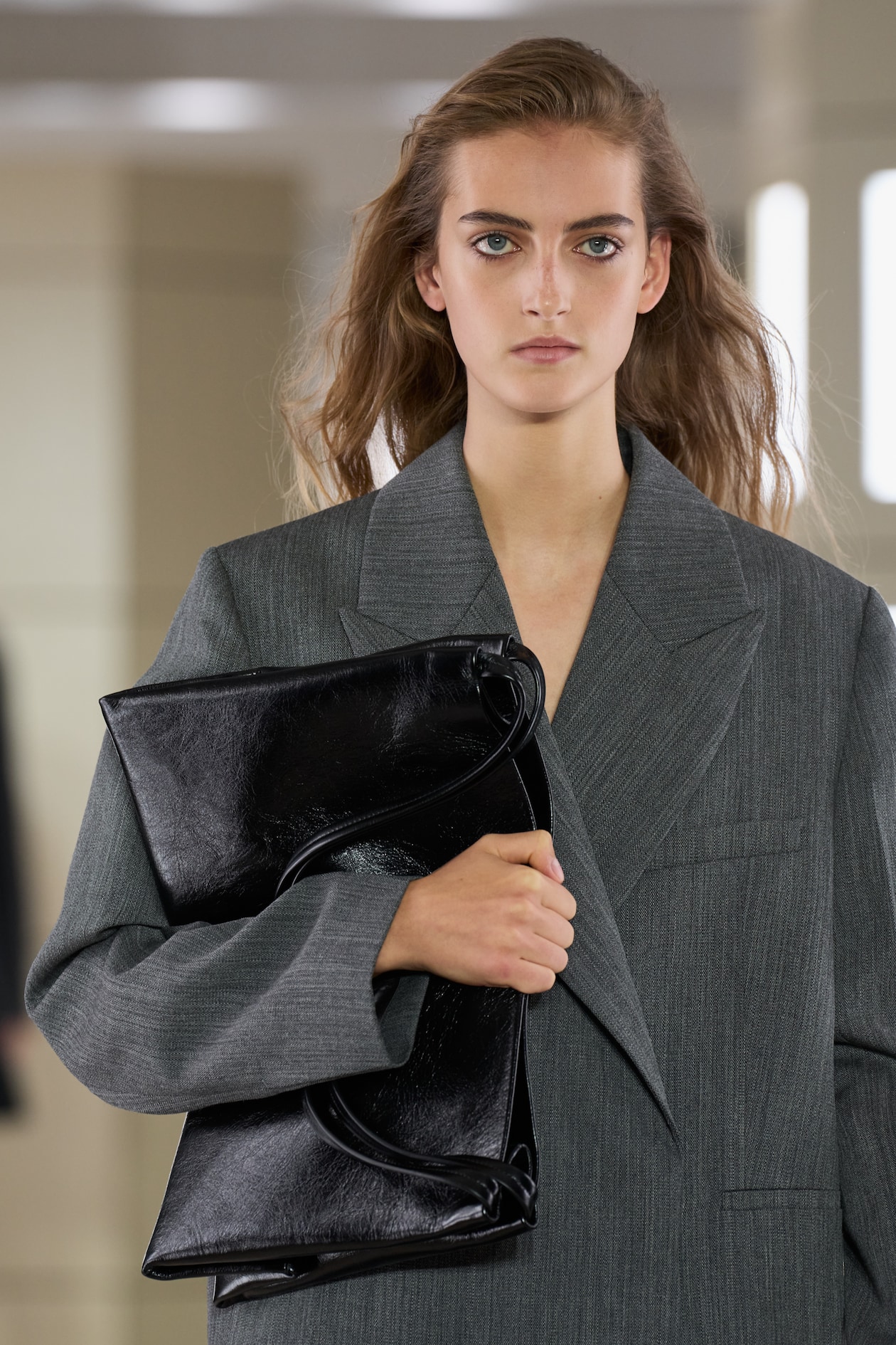 Person in a gray blazer holding a large black leather clutch, with long wavy hair and neutral expression against a blurred background.
