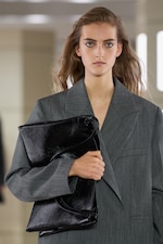 Placeholder for: Person in a gray blazer holding a large black leather clutch, with long wavy hair and neutral expression against a blurred background.