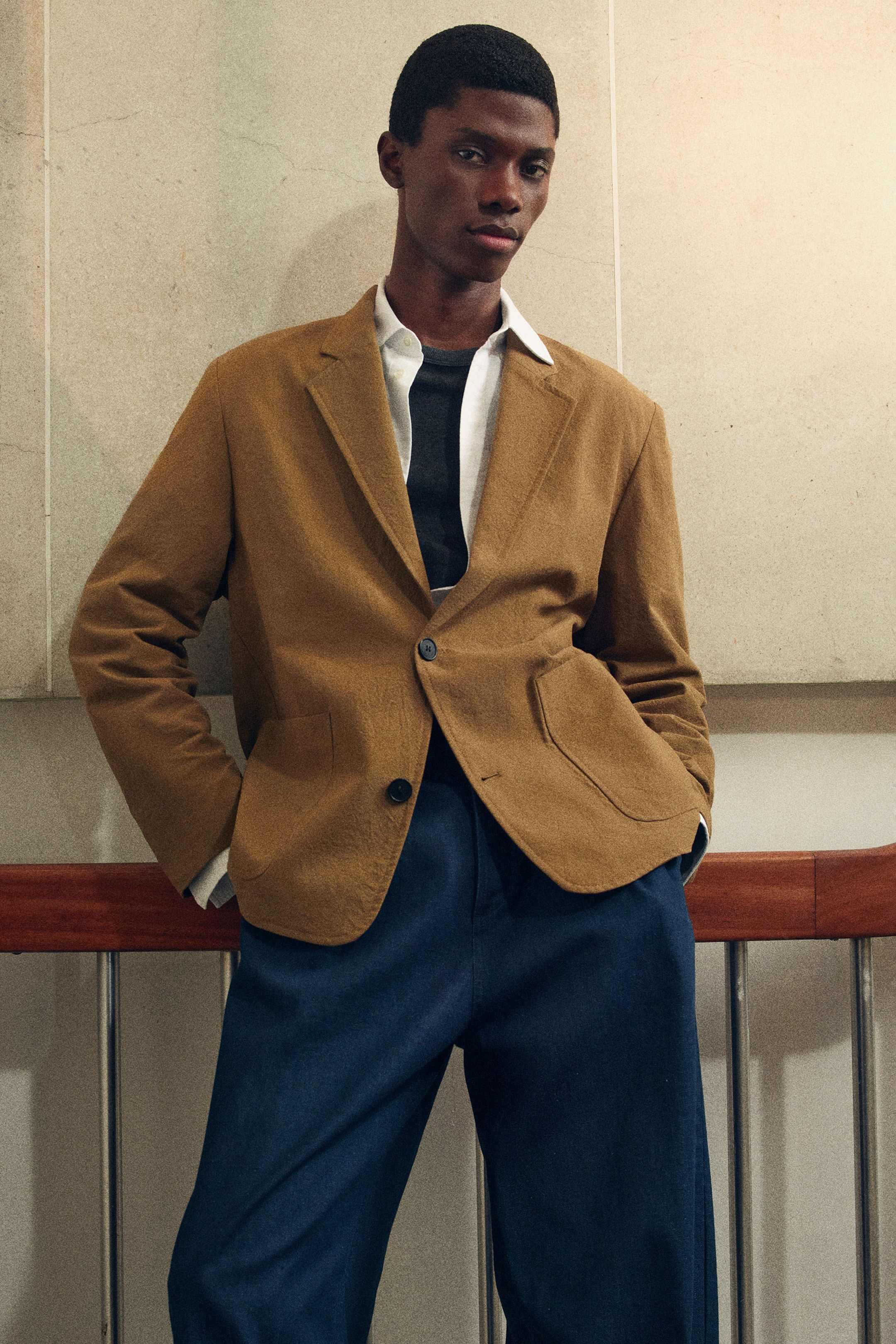 Man in a brown blazer and blue pants stands confidently, leaning against a railing with hands in pockets. Neutral wall background.