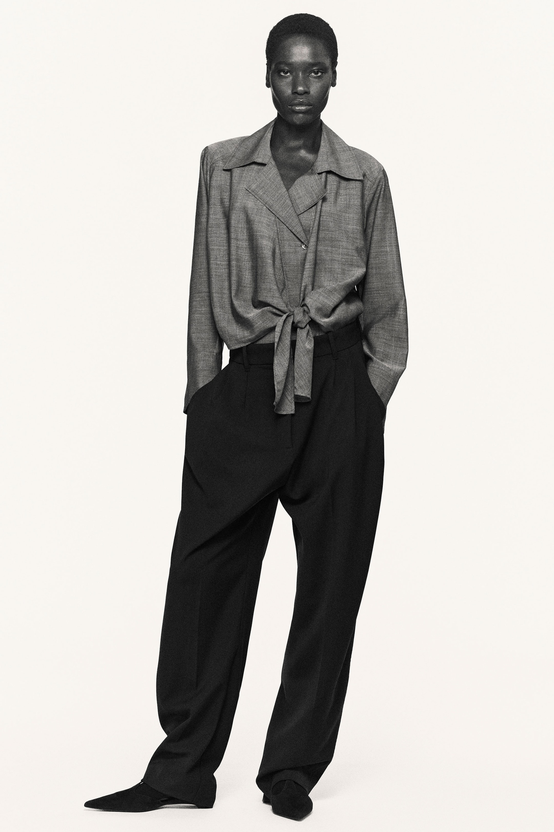Person in a buttoned shirt and wide-leg pants, standing with one hand in pocket. Monochrome image with a neutral background.
