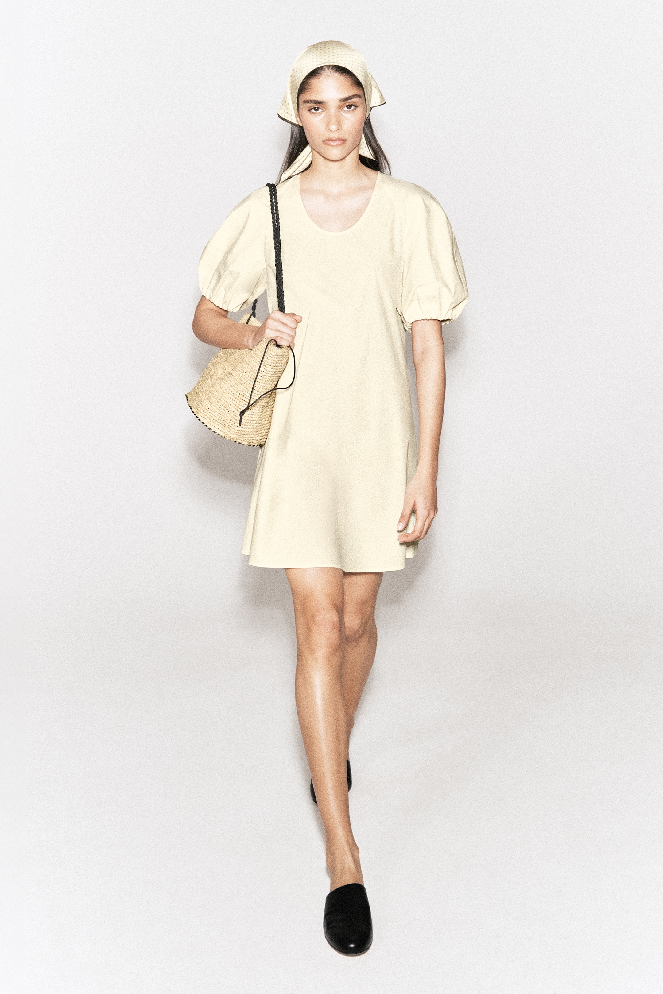 Woman in a beige dress and headscarf, carrying a straw bag, walks confidently in black shoes against a plain backdrop.