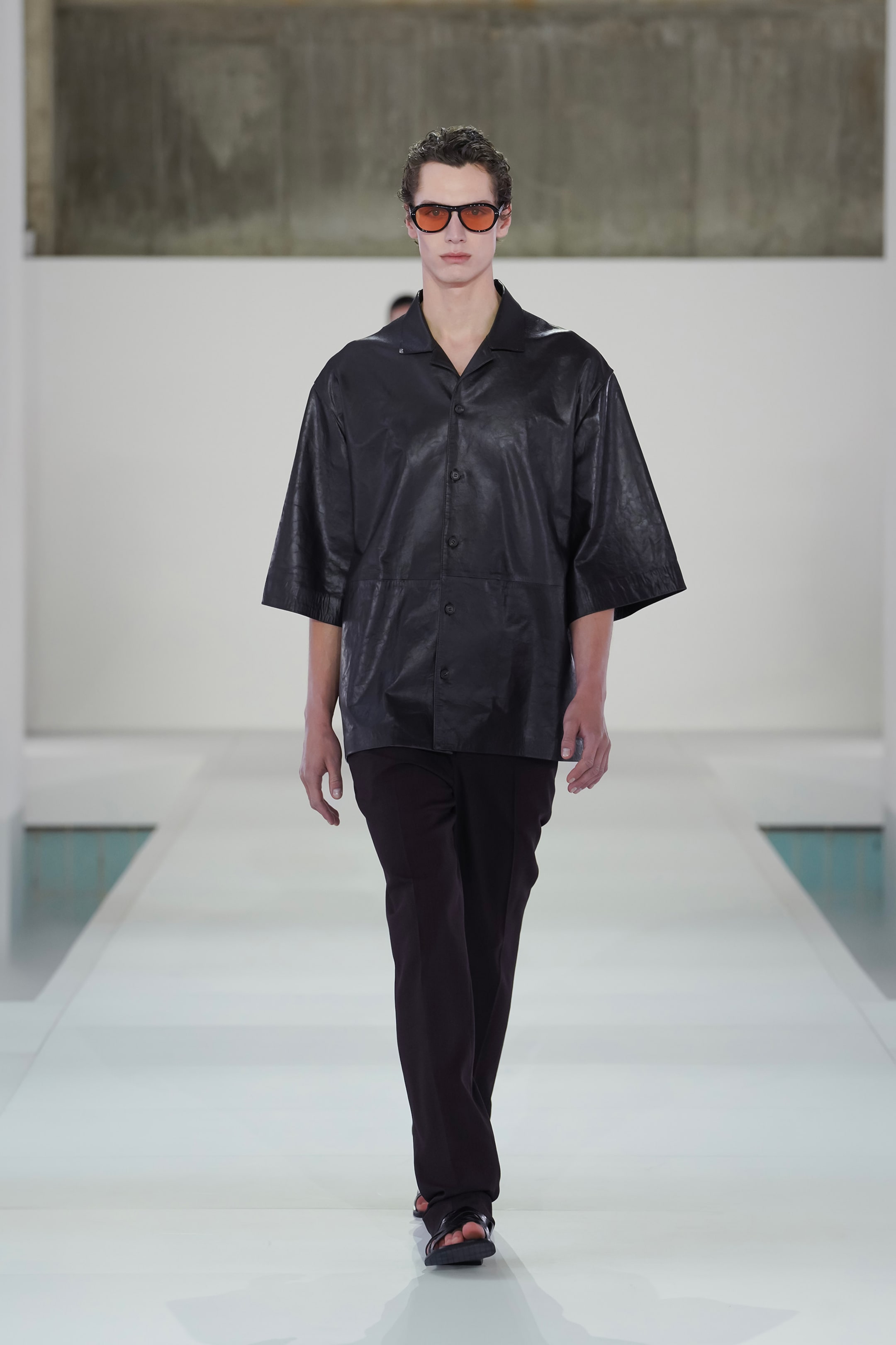 Model in black leather shirt and dark pants walks on a white runway, wearing sunglasses, against a minimalist background.
