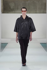 Placeholder for: Model in black leather shirt and dark pants walks on a white runway, wearing sunglasses, against a minimalist background.