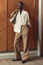 Placeholder for: A person poses confidently in a striped shirt, brown coat, and wide-legged pants against a wooden backdrop.