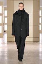 Placeholder for: Model wearing an oversized black suit with a matching scarf walks on a glossy floor with beige panel walls in the background.